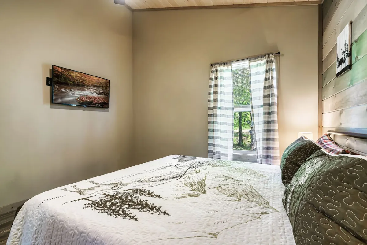 A tranquil master bedroom featuring a king-sized bed with a patterned quilt, a window with soft curtains letting in natural light, and a wall-mounted TV. The cozy setup invites relaxation and quiet moments, embodying a couple’s private retreat.