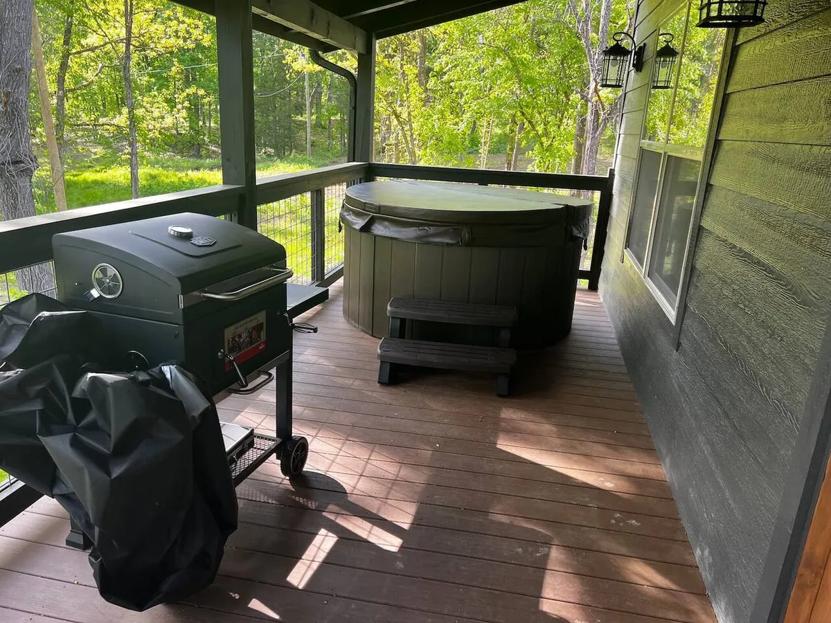 A broad view of a wooden deck features a grill, hot tub, and dining area, all nestled among tall trees. The deck invites guests to start their day with coffee in nature or enjoy a cozy evening outside, blending comfort with the beauty of the outdoors.