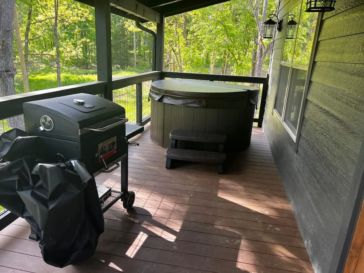 An inviting exterior deck area with a grill and hot tub, overlooking forested surroundings. The space is designed for barbecuing, relaxing in the hot tub, or simply enjoying a morning coffee while taking in the serene views. The natural backdrop suggests a seamless connection between the cabin and its outdoor environment.