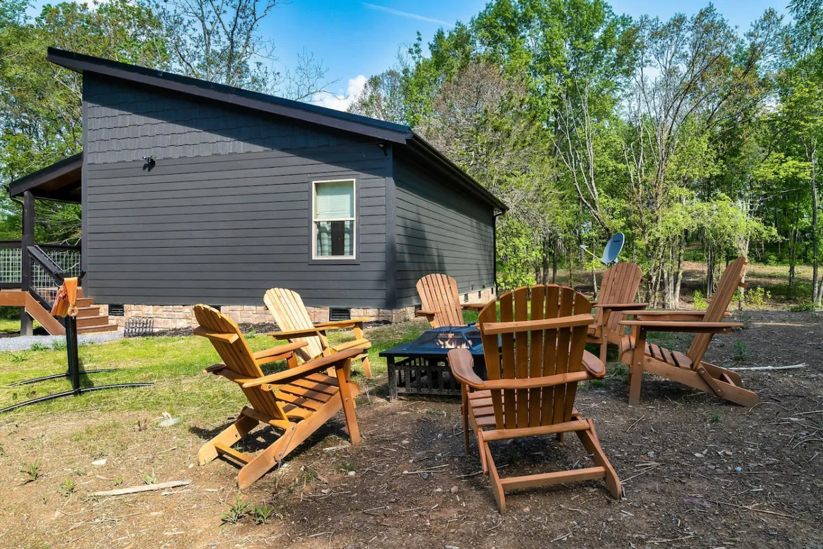 This exterior shot captures a circle of wooden Adirondack chairs around a fire pit, framed by natural surroundings. The setting is intimate and cozy, ideal for sharing stories, roasting marshmallows, or simply enjoying the sights and sounds of the forest.