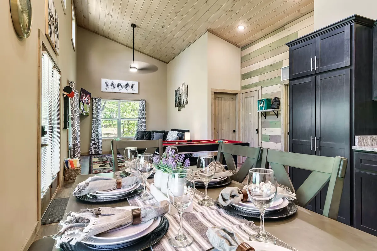 A welcoming dining area with a table set for six, illuminated by natural light streaming through large windows. Decorative wall art adds personality, and the overall mood is cozy, perfect for family dinners, game nights, or celebrations with friends.