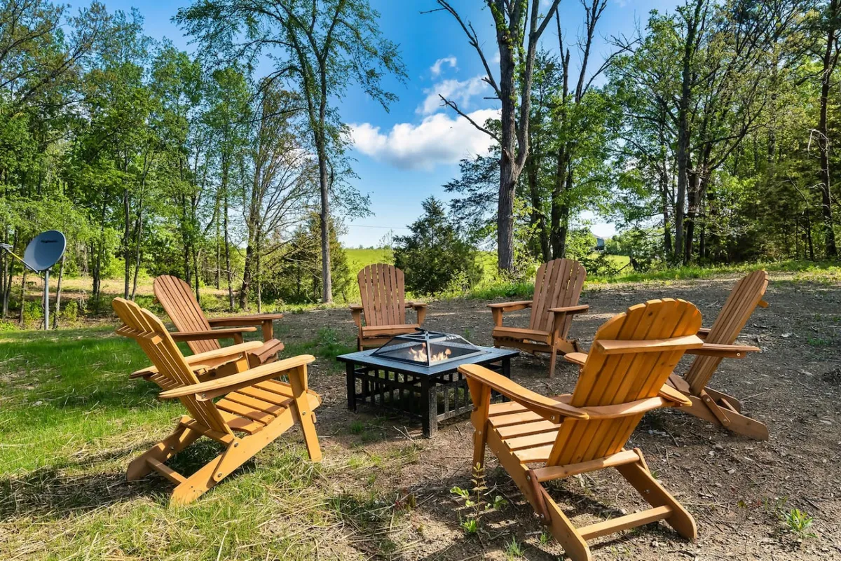 This exterior image features a cozy arrangement of Adirondack chairs encircling a fire pit, set amidst lush surrounding greenery. The setting is open to the sky, perfect for both sunset viewing and nighttime stargazing. The natural landscape and seating invite outdoor gatherings and moments of relaxation in nature.