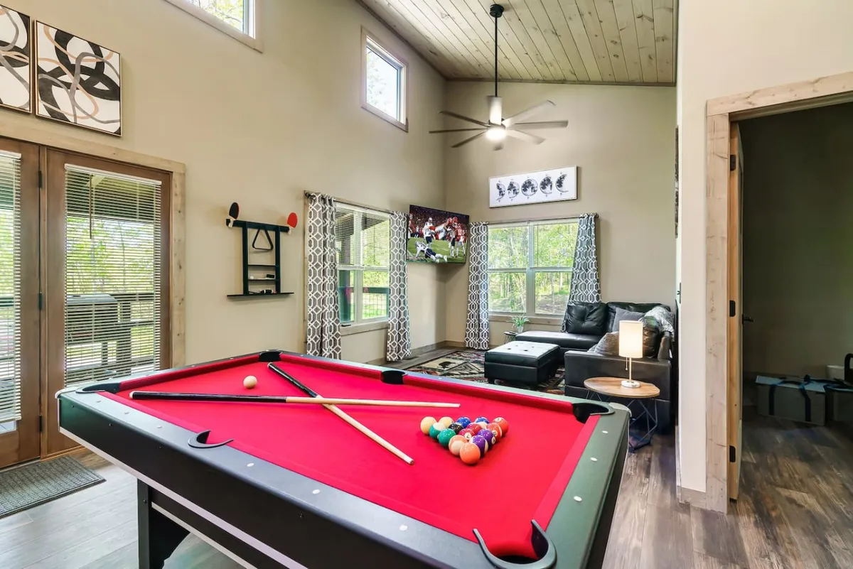 A vibrant living and entertainment area featuring a pool table, large windows framing tranquil woods, a comfortable sofa facing a big-screen television, and an inviting, open atmosphere. Sunlight streams in, enhancing the cheerful and lively mood of the space.