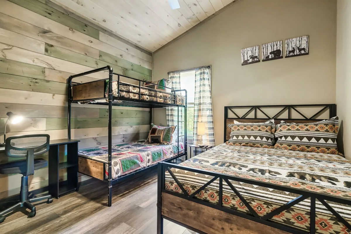 A spacious second bedroom with a queen bed, a twin-over-twin bunk, a desk with chair for work or study, and a window draped with curtains. The scene feels relaxed and family-friendly, ideal for both sleep and productivity.