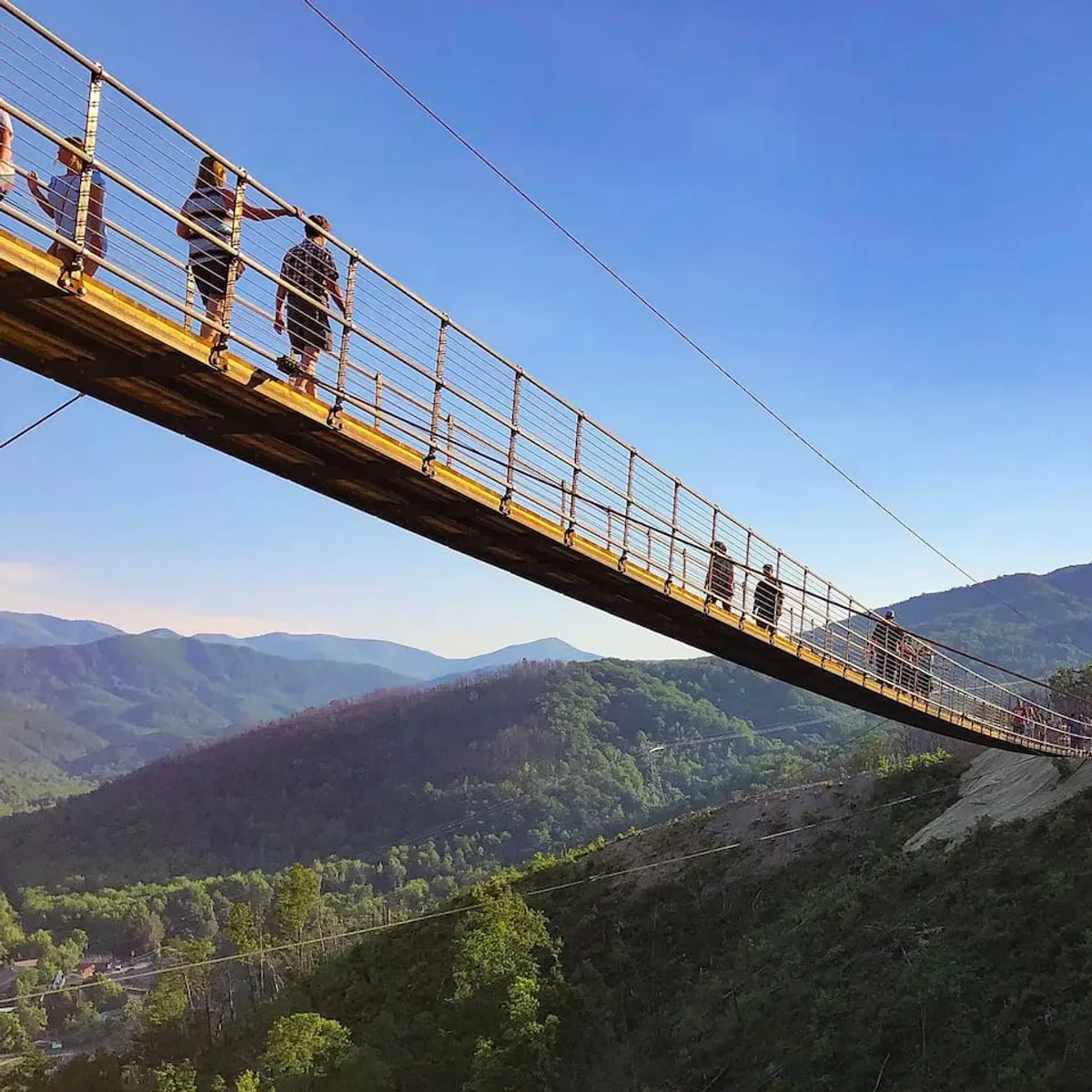 An exterior shot highlighting a dramatic suspension bridge spanning a mountain landscape, with people trekking across and breathtaking views stretching into the distance. The image evokes the thrill of outdoor adventure unique to the Smoky Mountains.
