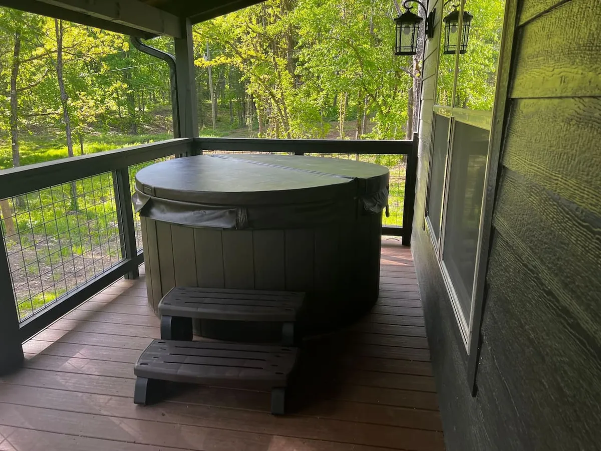 This image showcases a hot tub on a wooden deck surrounded by lush greenery and softly glowing lanterns. The natural setting, combined with the inviting hot tub, creates an atmosphere perfect for relaxing under the stars, enjoying nature, or sharing a romantic moment with a partner.