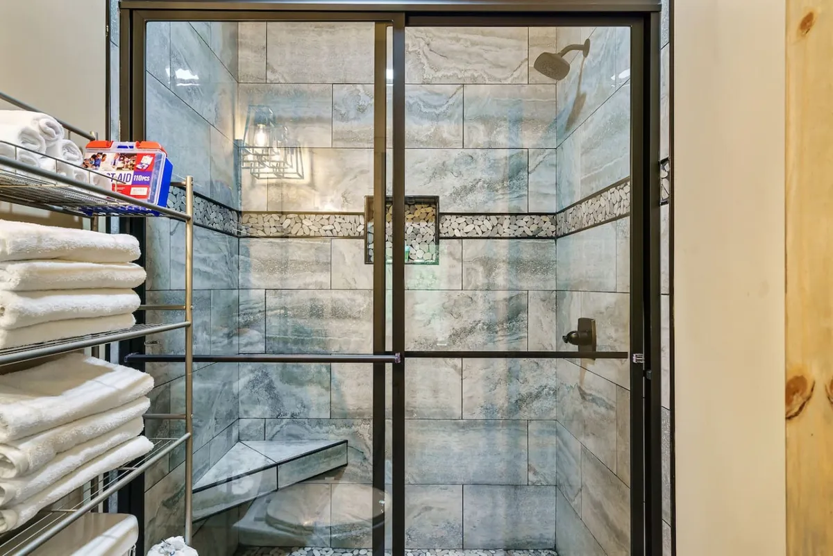 A refreshing bathroom with a glass-enclosed walk-in shower, textured stone tiles, a neat shelf of stacked towels, and a decorative light fixture. The composition feels spa-inspired and tranquil.